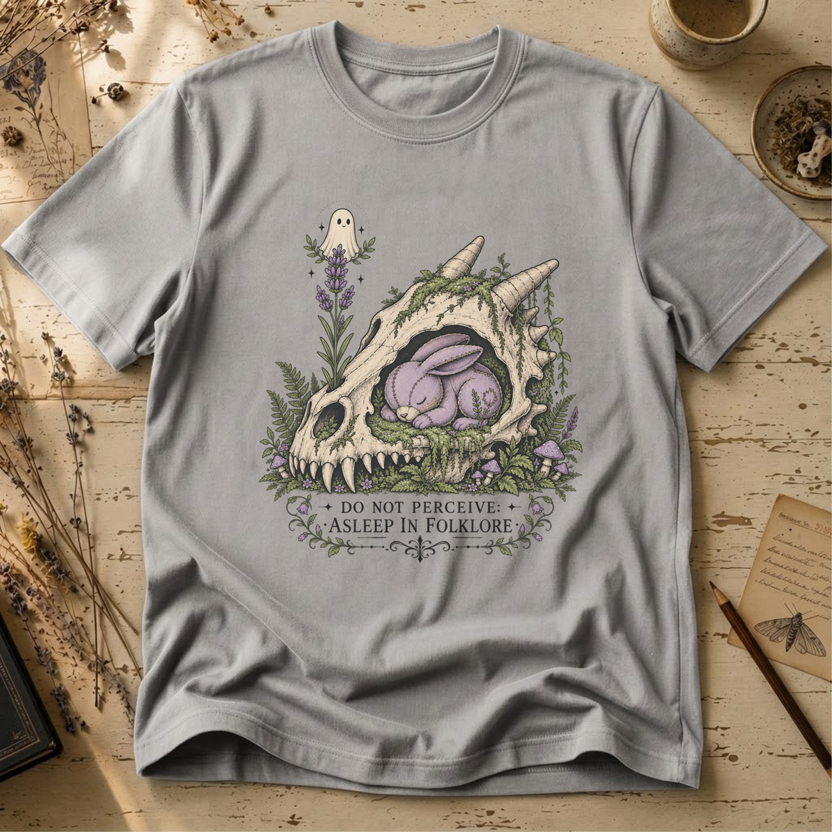 Light gray t-shirt with an illustration of a horned, fanged skull overgrown with moss and lavender, a sleeping purple stitched rabbit curled inside it, and a small ghost hovering above.