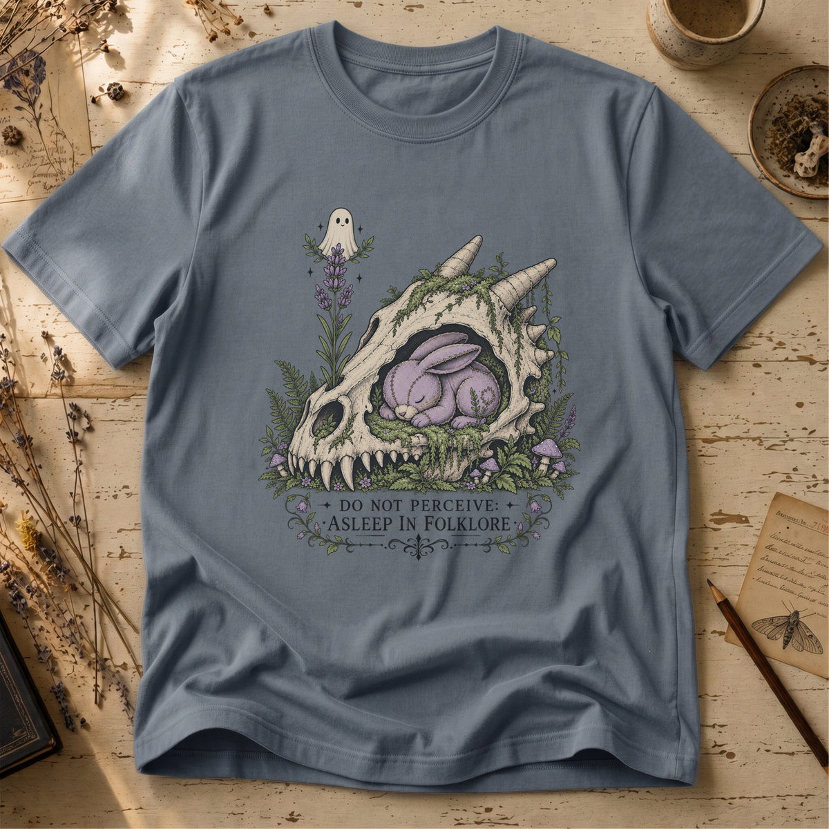 Steel blue t-shirt with an illustration of a horned, fanged skull overgrown with moss and lavender, a sleeping purple stitched rabbit curled inside it, and a small ghost hovering above.