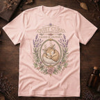 Soft pink t-shirt with a vintage apothecary label design: a sleeping fawn with white spots framed in an oval of lavender sprigs and a pink rose, labeled 'Elixir of Solitude'.