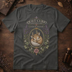 Dark gray t-shirt with a vintage apothecary label design: a sleeping fawn with white spots framed in an oval of lavender sprigs and a pink rose, labeled 'Elixir of Solitude'.