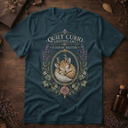 Deep teal t-shirt with a vintage apothecary label design: a sleeping fawn with white spots framed in an oval of lavender sprigs and a pink rose, labeled 'Elixir of Solitude'.