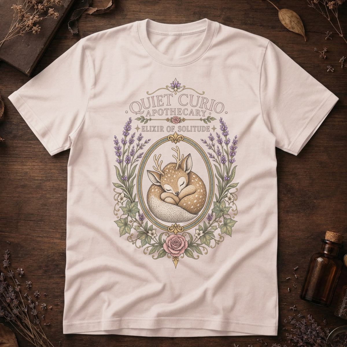 Vintage white t-shirt with a vintage apothecary label design: a sleeping fawn with white spots framed in an oval of lavender sprigs and a pink rose, labeled 'Elixir of Solitude'.