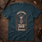 Deep teal t-shirt with a gothic herbarium illustration: a skull-shaped plant sprouting purple and pink flowers above an open botanical book, framed in an arch with a crescent moon.