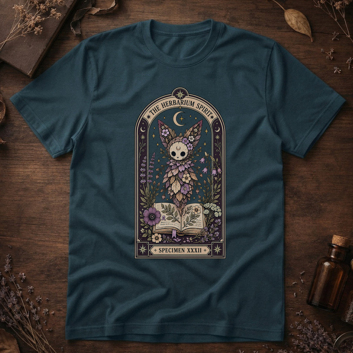 Deep teal t-shirt with a gothic herbarium illustration: a skull-shaped plant sprouting purple and pink flowers above an open botanical book, framed in an arch with a crescent moon.