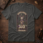 Charcoal gray t-shirt with a gothic herbarium illustration: a skull-shaped plant sprouting purple and pink flowers above an open botanical book, framed in an arch with a crescent moon.