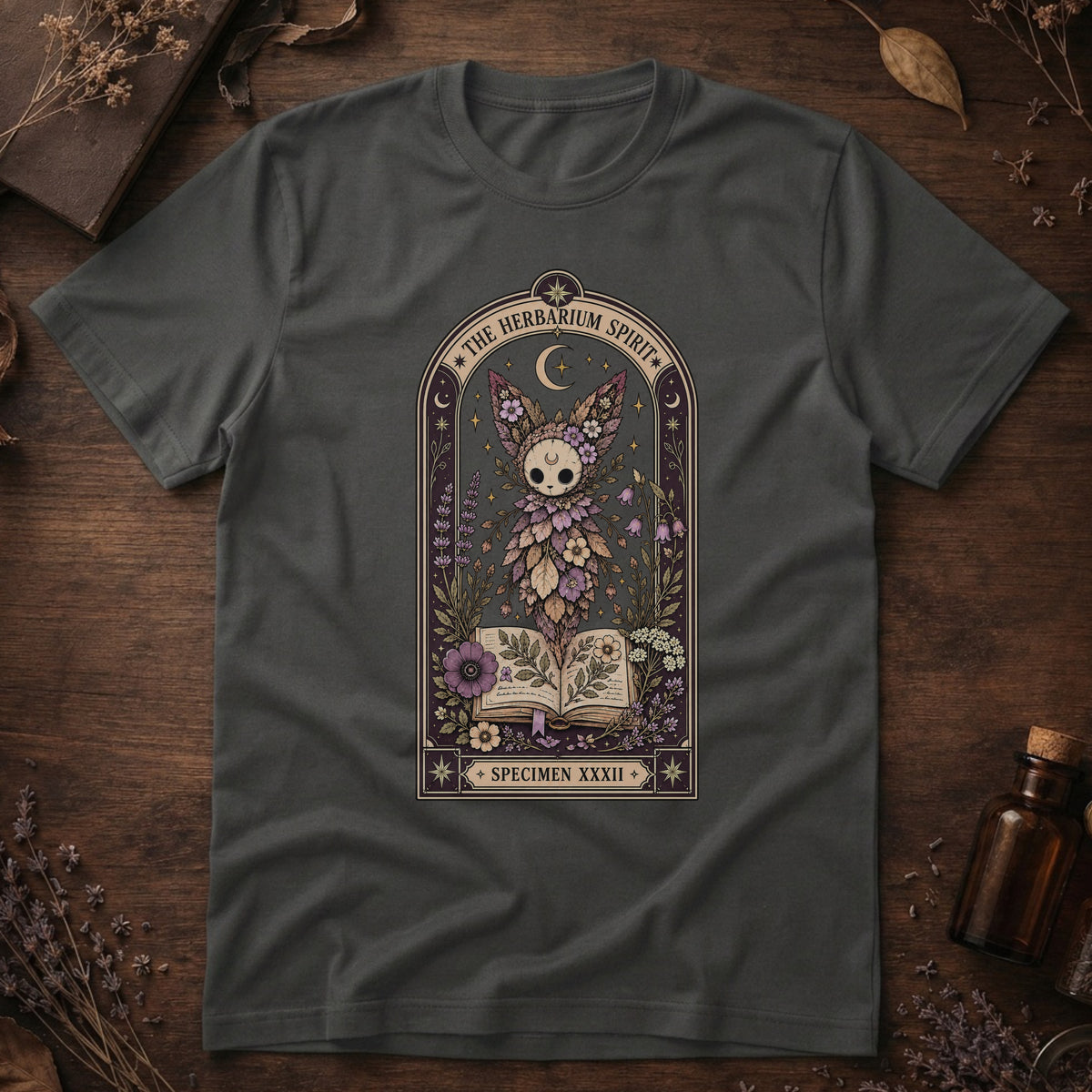 Charcoal gray t-shirt with a gothic herbarium illustration: a skull-shaped plant sprouting purple and pink flowers above an open botanical book, framed in an arch with a crescent moon.