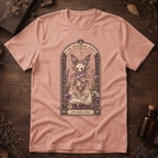 Peach t-shirt with a gothic herbarium illustration: a skull-shaped plant sprouting purple and pink flowers above an open botanical book, framed in an arch with a crescent moon.