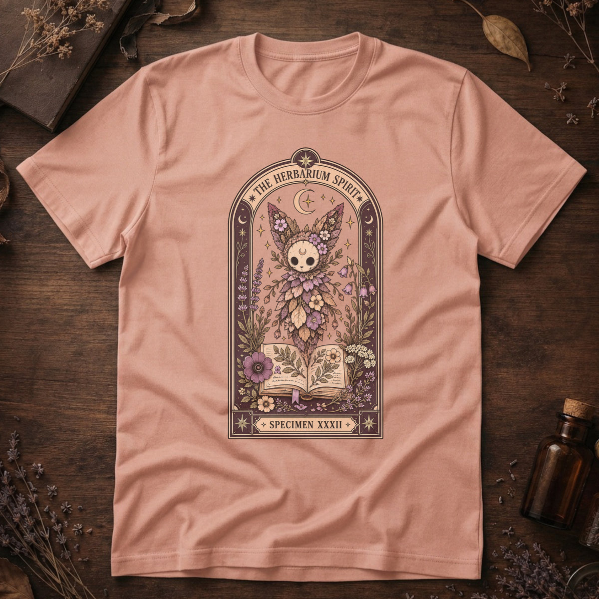 Peach t-shirt with a gothic herbarium illustration: a skull-shaped plant sprouting purple and pink flowers above an open botanical book, framed in an arch with a crescent moon.