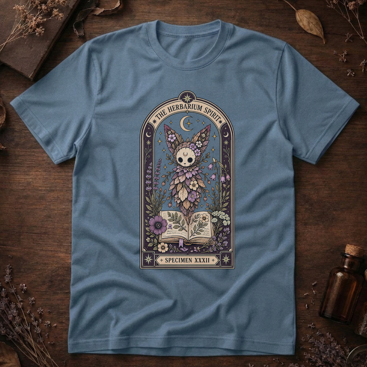 Steel blue t-shirt with a gothic herbarium illustration: a skull-shaped plant sprouting purple and pink flowers above an open botanical book, framed in an arch with a crescent moon.