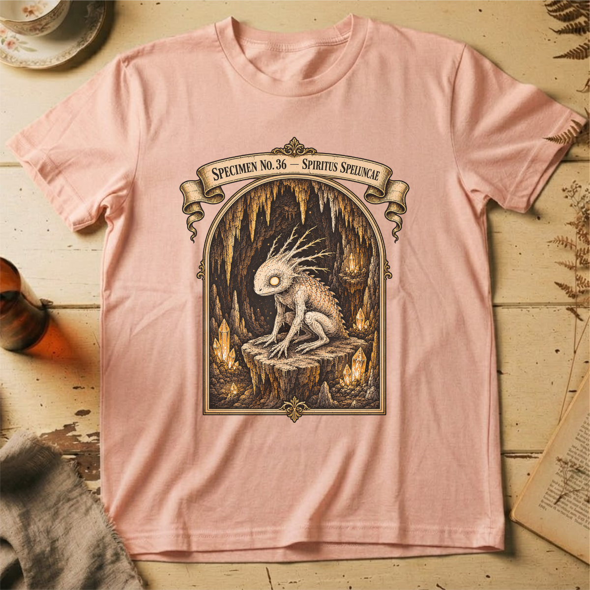 Peach t-shirt with a gothic illustration of a horned, single-eyed cave spirit perched on glowing crystal formations inside an arched cavern frame.