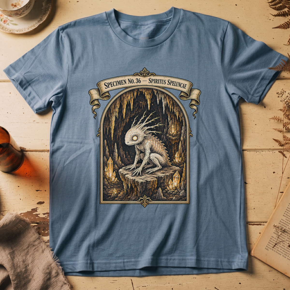 Steel blue t-shirt with a gothic illustration of a horned, single-eyed cave spirit perched on glowing crystal formations inside an arched cavern frame.