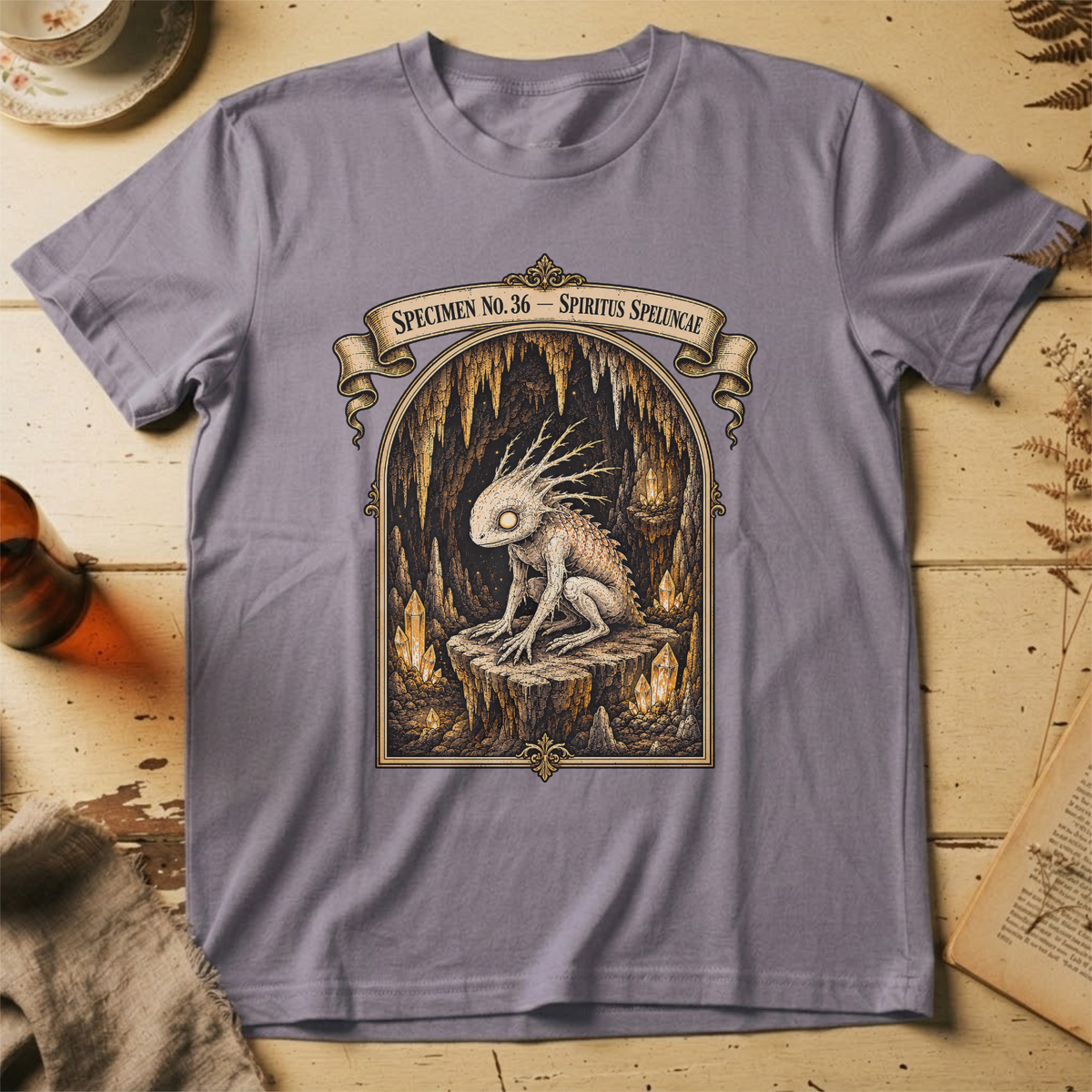 Storm grey t-shirt with a gothic illustration of a horned, single-eyed cave spirit perched on glowing crystal formations inside an arched cavern frame.
