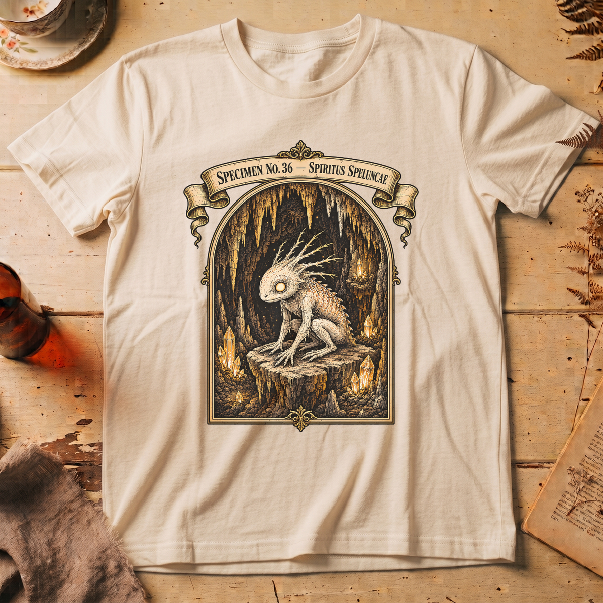 Vintage white t-shirt with a gothic illustration of a horned, single-eyed cave spirit perched on glowing crystal formations inside an arched cavern frame.
