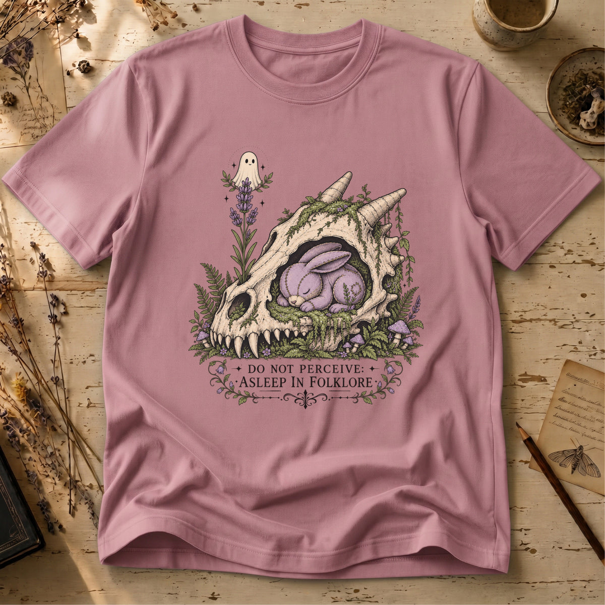 Orchid pink t-shirt with an illustration of a horned, fanged skull overgrown with moss and lavender, a sleeping purple stitched rabbit curled inside it, and a small ghost hovering above.