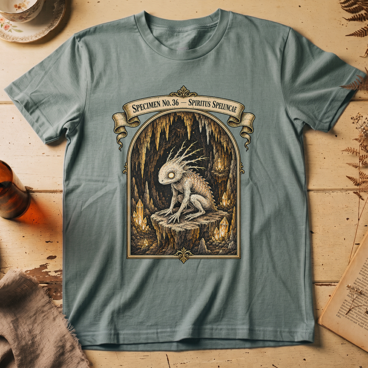 Sage green t-shirt with a gothic illustration of a horned, single-eyed cave spirit perched on glowing crystal formations inside an arched cavern frame.