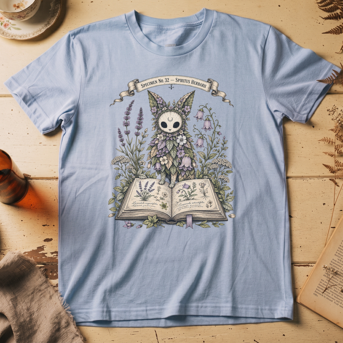 Baby blue t-shirt with a botanical illustration of a skull-faced spirit wearing a floral crown, standing among lavender and bluebell flowers between two open books.