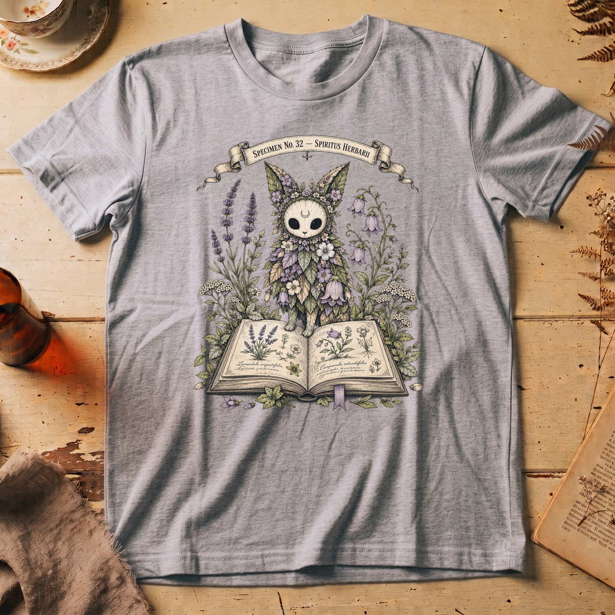 Silver t-shirt with a botanical illustration of a skull-faced spirit wearing a floral crown, standing among lavender and bluebell flowers between two open books.