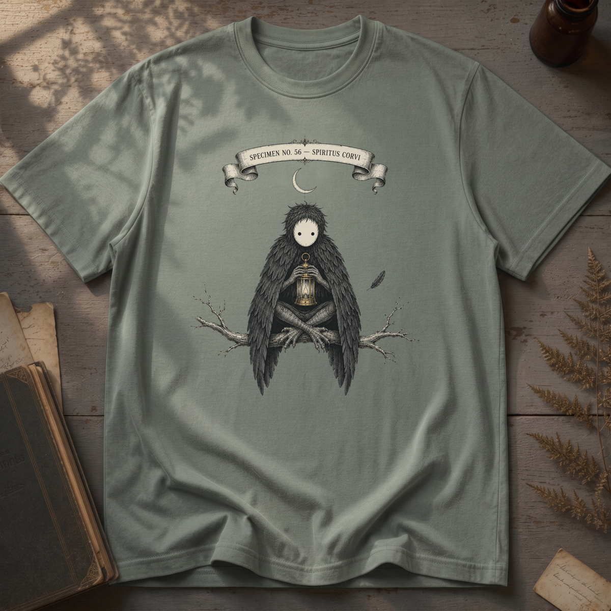 Sage green t-shirt with a gothic illustration of a raven-winged spirit sitting on a bare branch, holding a glowing lantern, beneath a crescent moon banner reading Specimen No. 56.