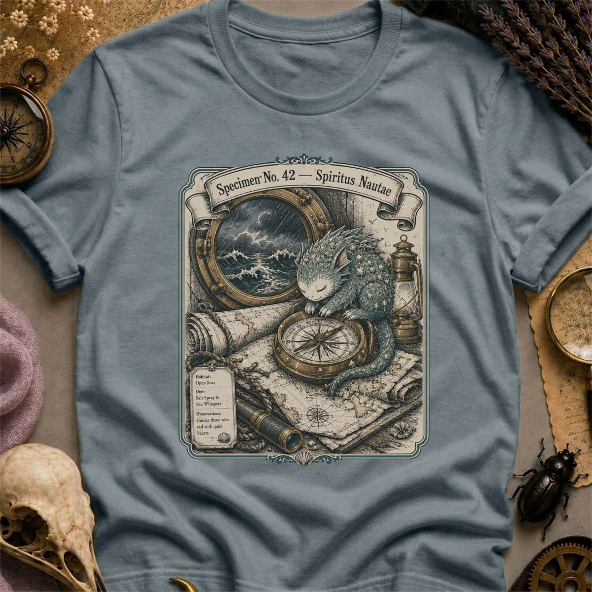 Steel blue t-shirt with a sepia-toned illustration of a sea spirit resting a paw on a brass compass atop an antique nautical map, with a stormy porthole scene above.