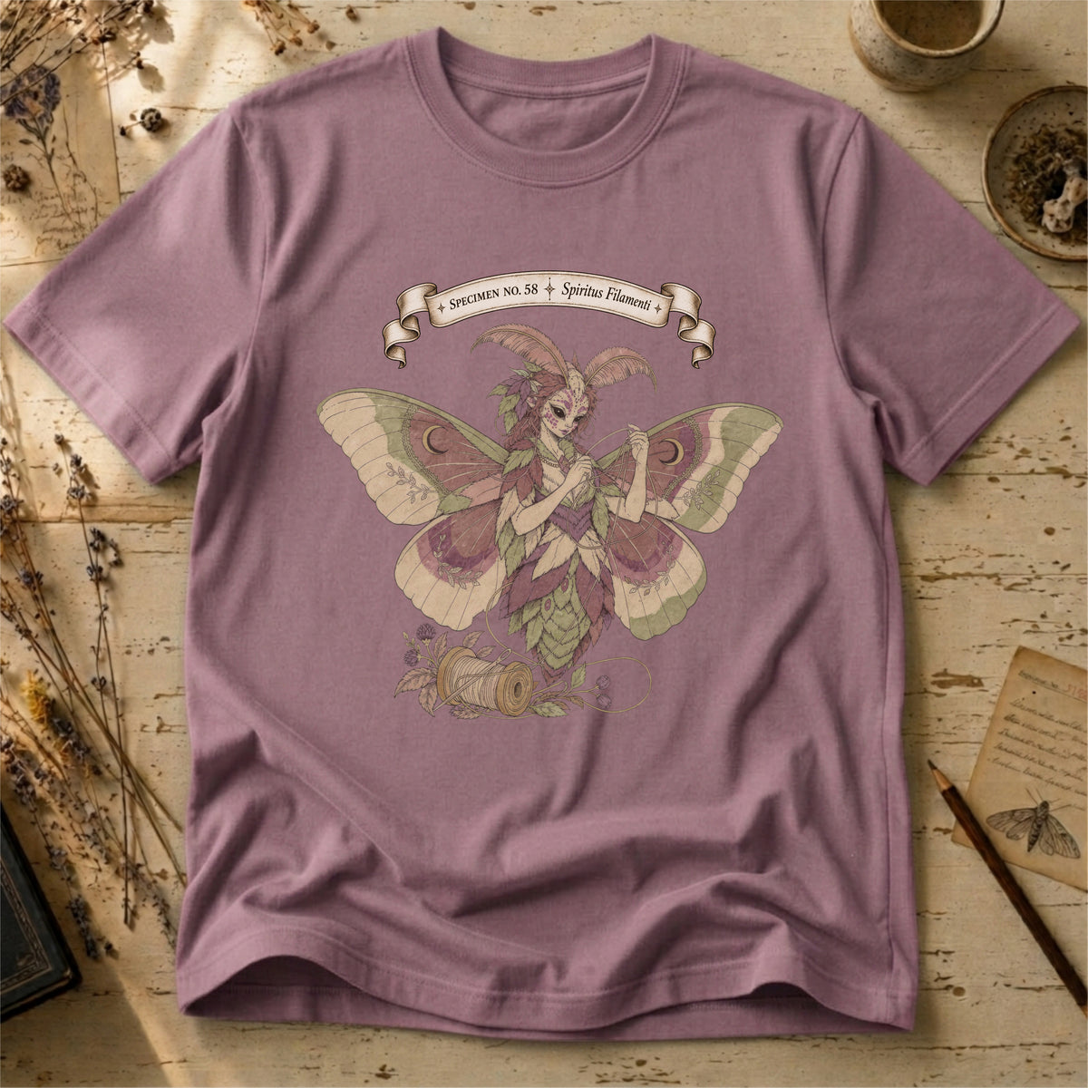 Orchid pink t-shirt with an illustration of a moth-winged spirit holding a strand of gold thread beside a spool and needle, surrounded by dusty pink flowers and sage leaves, beneath a banner reading Specimen No. 58.