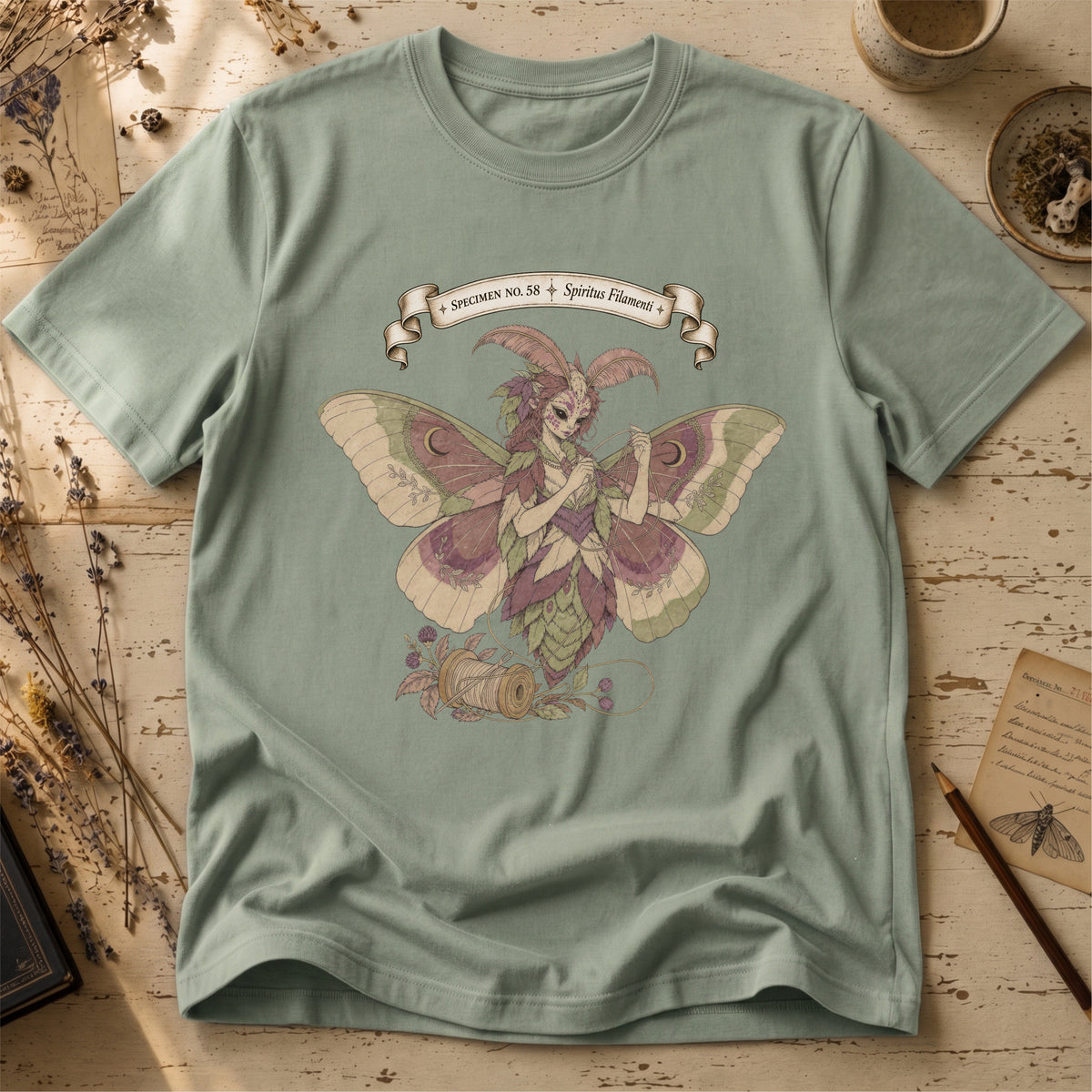 Sage green t-shirt with an illustration of a moth-winged spirit holding a strand of gold thread beside a spool and needle, surrounded by dusty pink flowers and sage leaves, beneath a banner reading Specimen No. 58.