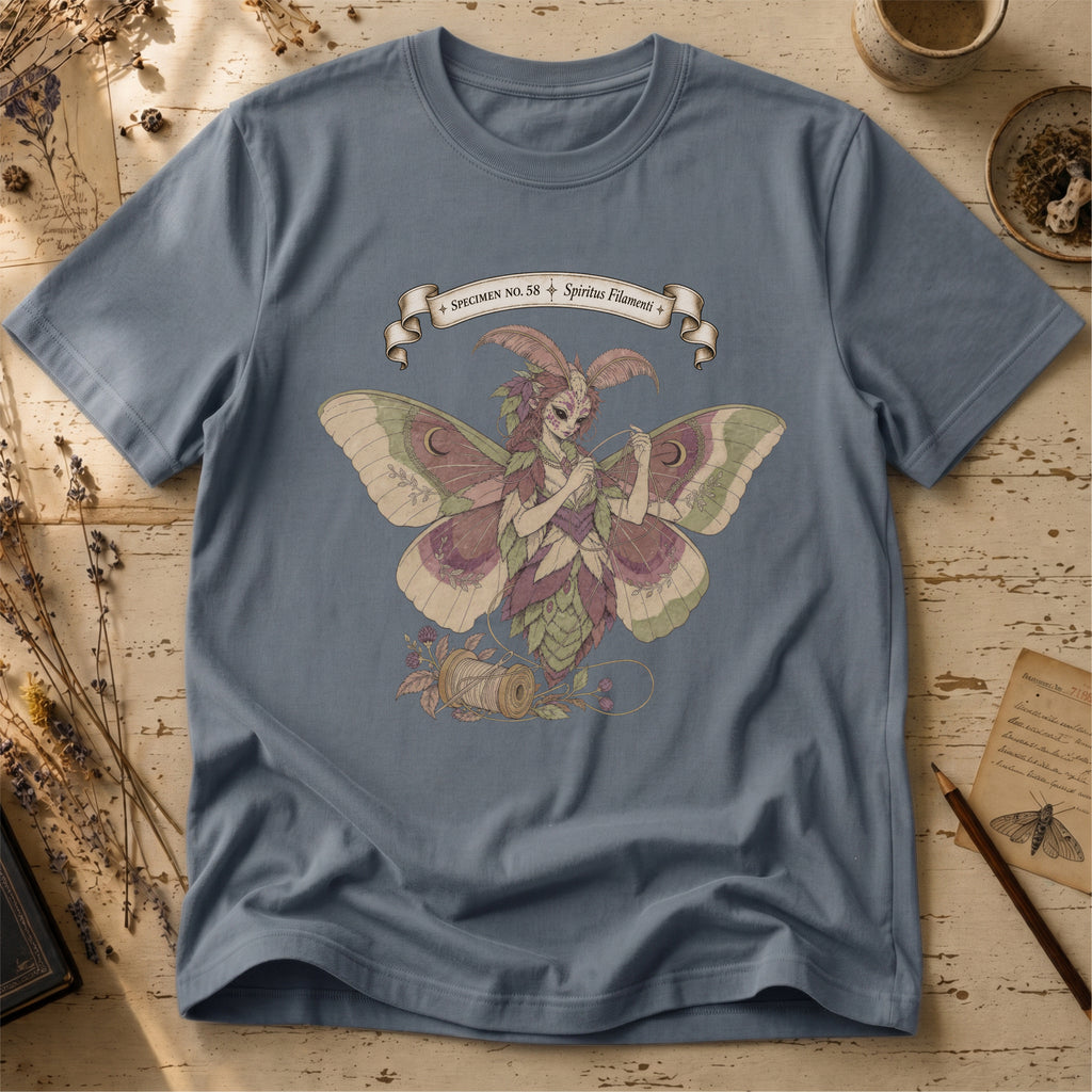 Steel blue t-shirt with an illustration of a moth-winged spirit holding a strand of gold thread beside a spool and needle, surrounded by dusty pink flowers and sage leaves, beneath a banner reading Specimen No. 58.