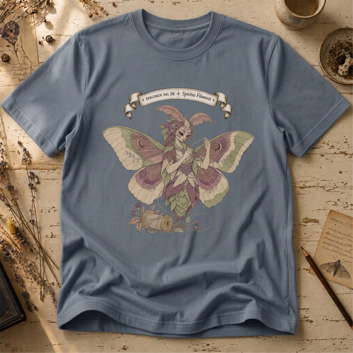 Steel blue t-shirt with an illustration of a moth-winged spirit holding a strand of gold thread beside a spool and needle, surrounded by dusty pink flowers and sage leaves, beneath a banner reading Specimen No. 58.