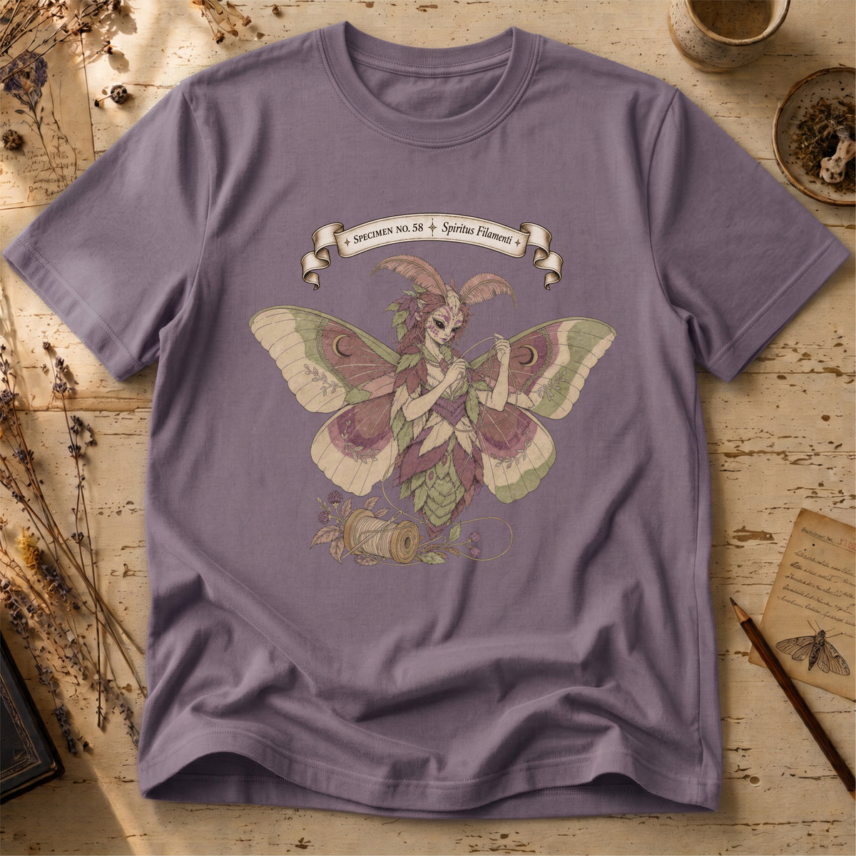 Storm grey t-shirt with an illustration of a moth-winged spirit holding a strand of gold thread beside a spool and needle, surrounded by dusty pink flowers and sage leaves, beneath a banner reading Specimen No. 58.