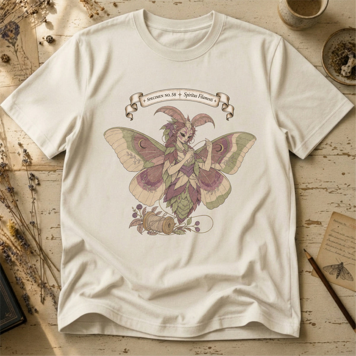 Vintage white t-shirt with an illustration of a moth-winged spirit holding a strand of gold thread beside a spool and needle, surrounded by dusty pink flowers and sage leaves, beneath a banner reading Specimen No. 58.